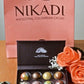 Caja de regalo negra mate con 8 bombones artesanales pintados a mano de NIKADI, elaborados con cacao ancestral colombiano de origen "Bean to Bonbon". Incluye bombones metalizados y jaspeados, presentados junto a una bolsa rosa NIKADI y rosas melocotón, exclusivo en Casa Precis, Concept Store.