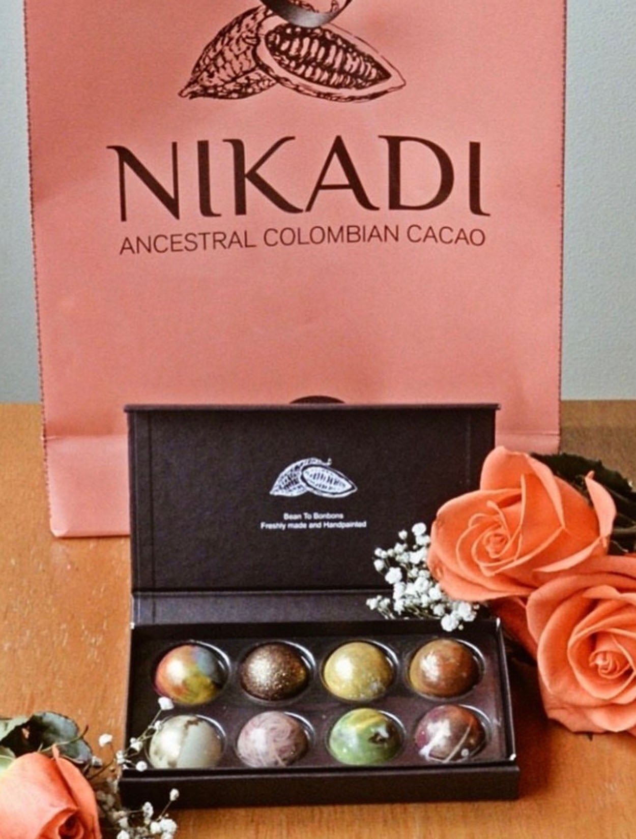 Caja de regalo negra mate con 8 bombones artesanales pintados a mano de NIKADI, elaborados con cacao ancestral colombiano de origen "Bean to Bonbon". Incluye bombones metalizados y jaspeados, presentados junto a una bolsa rosa NIKADI y rosas melocotón, exclusivo en Casa Precis, Concept Store.