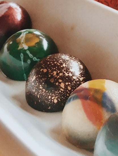 Caja de regalo negra mate con 8 bombones artesanales pintados a mano de NIKADI, elaborados con cacao ancestral colombiano de origen "Bean to Bonbon". Incluye bombones metalizados y jaspeados, presentados junto a una bolsa rosa NIKADI y rosas melocotón, exclusivo en Casa Precis, Concept Store.
