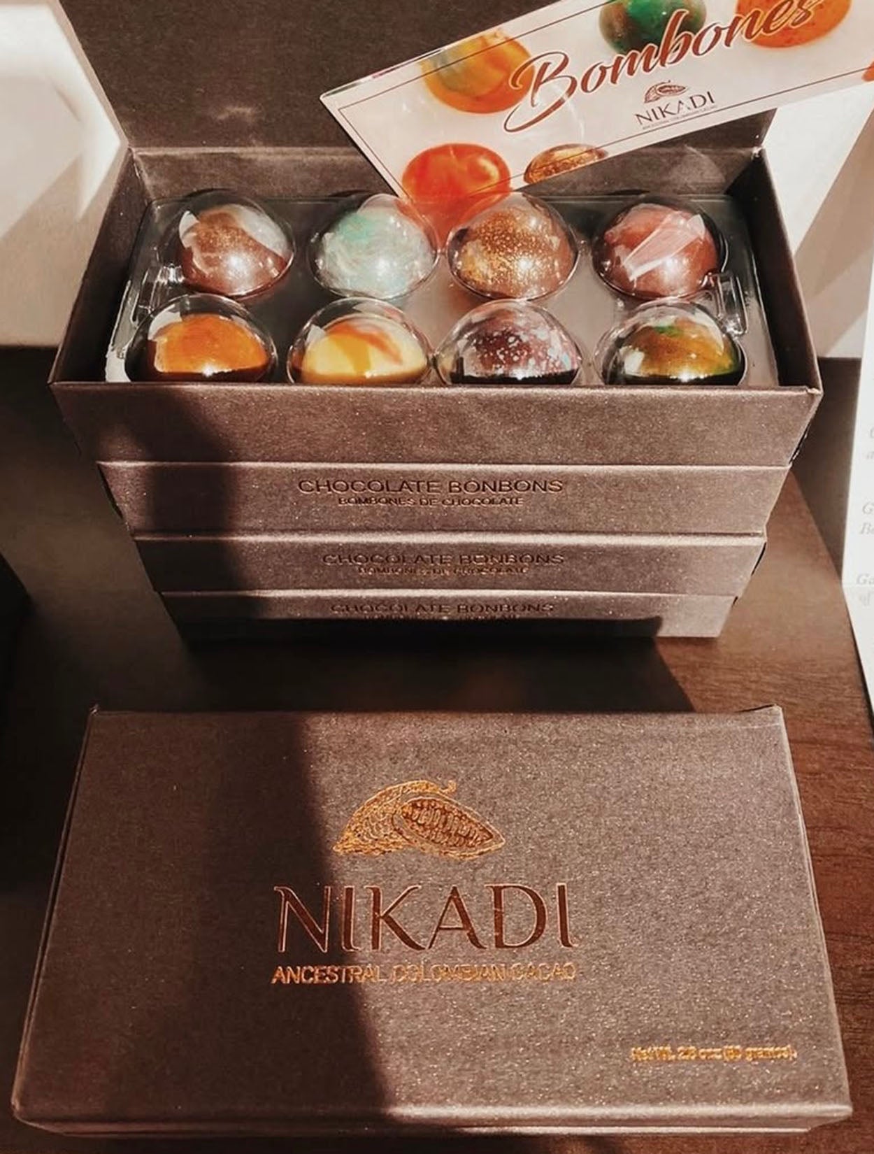 Caja de regalo negra mate con 8 bombones artesanales pintados a mano de NIKADI, elaborados con cacao ancestral colombiano de origen "Bean to Bonbon". Incluye bombones metalizados y jaspeados, presentados junto a una bolsa rosa NIKADI y rosas melocotón, exclusivo en Casa Precis, Concept Store.